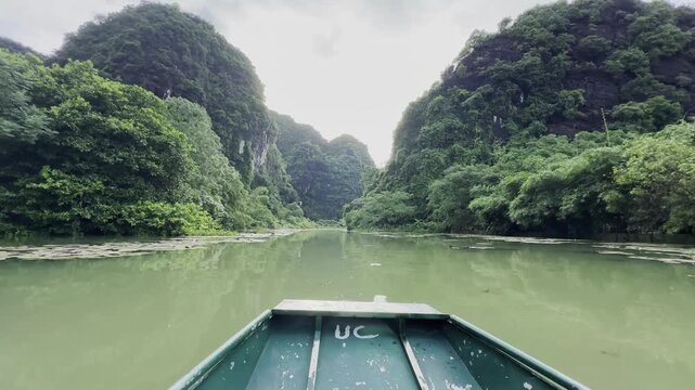 A slow boat ride along a narrow emerald river in Tam Coc, Vietnam, passing water lilies, lotus flowers, lush green vegetation and dramatic karst mountains, creating a calm nature experience.