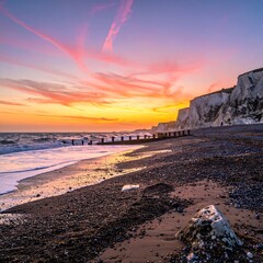 Sunrise over a pebbled beach with cliffs, vibrant hues of orange and pink painting the sky, waves gently crash