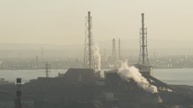 Industrial factory smokestacks emitting white smoke into a hazy sky.