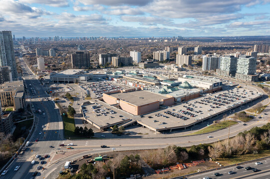 Aerial view capturing a large urban shopping mall surrounded by multi level parking lots and busy roads, with residential high rise buildings and toronto's skyline in the distance