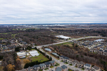 Fototapeta premium Sprawling residential neighborhood featuring modern houses, a network of roads, and green spaces, bordered by natural treed areas and distant urban infrastructure under an overcast sky