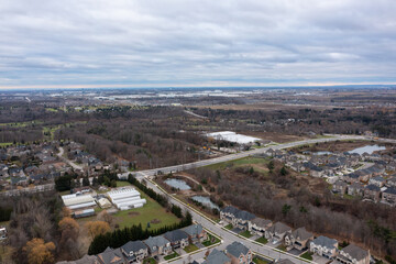 Residential houses and quiet streets wind through developing suburban landscape, featuring bare trees and a cloudy sky, with industrial buildings visible in the distance