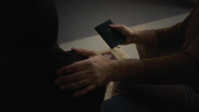 Over the shoulder view of unrecognisable religious mature man holding bible book supporting anonymous crying woman