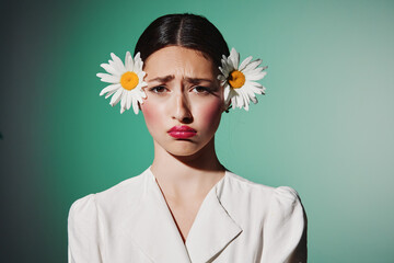 Young woman with two daisies in her hair wears white clothing and pouty lips against a vibrant green background. Concept of fashion, beauty creativity, and tender emotions.