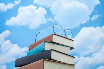 Stack of books with eyeglasses against a blue sky with clouds. Education, knowledge, learning, academic growth concept.