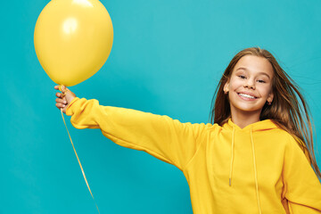 Smiling girl in yellow hoodie holding yellow balloon with outstretched arm on blue background,...