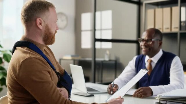 Caucasian man with injured arm looking through document while smiling senior Black insurance agent consulting him