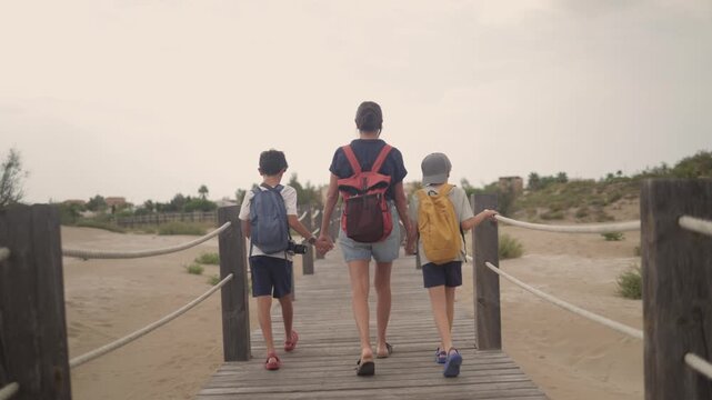 Family walking on a wooden boardwalk, mother and two children with backpacks exploring sandy dunes, enjoying outdoor summer travel, holiday, and togetherness on a beach path