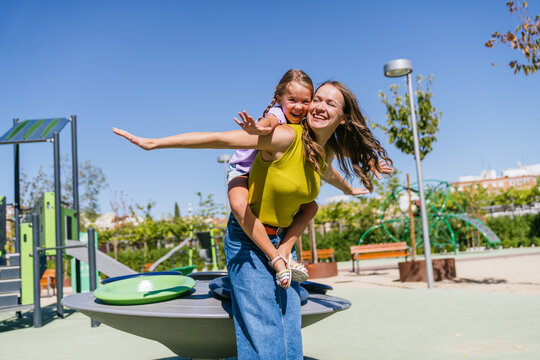 Young mother giving her happy daughter a piggyback ride on a sunny day in an urban playground, enjoying family fun