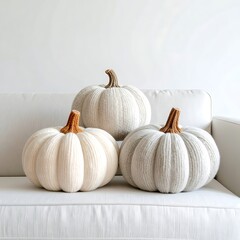 3 plush, knit pumpkins, in graduated shades of cream, off-white, and gray, on a white sofa against a white wall