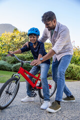 Fototapeta premium Asian father and son practicing riding red bicycle on gravel pathway in blue helmet