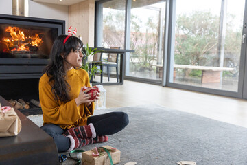 Naklejka premium Woman sitting cross-legged on rug in living room holding mug among wrapped gifts and fireplace