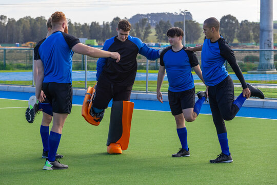 Diverse male hockey players linking arms, stretching quadriceps on turf in orange goalie pads