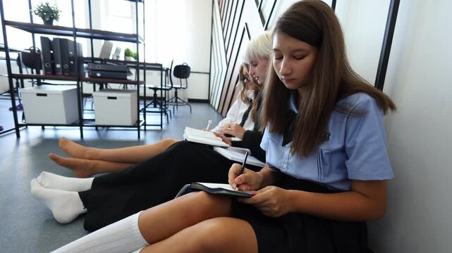 Three students sit on the floor, deeply focused on writing in notebooks, capturing a moment of quiet study and academic dedication in a bright, modern space.