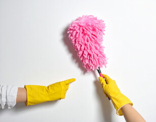 Two yellow gloved hands one holding a pink duster and the other pointing at it on a white wall