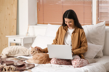 Pretty young woman with laptop and cute Maine Coon cat sitting on bed at home