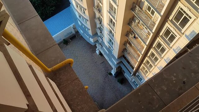 View down from top floor of multi-story building. Facade of white multi-story high-rise building with windows and balconies illuminated by light of setting sun and floor area underneath paved tiles