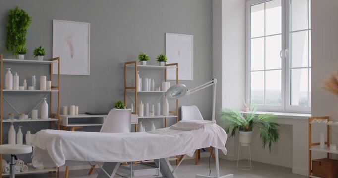 Interior of spa cosmetology clinic interior with massage table, shelves of cosmetics, green plants, and an adjustable lamp beside a large window. Calm, welcoming treatment space for clients concept