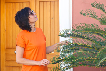 An adult woman a short bright dress in the garden of her house near a palm tree.