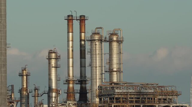 Industrial smokestacks and refinery infrastructure against a pale sky.