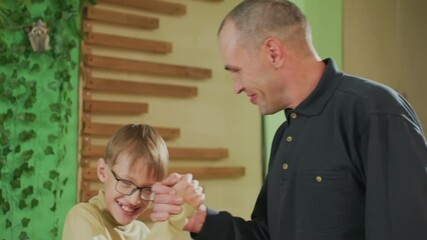 white father and son smiling at home, father praises sons drawing and offers handshake, cozy interior with green foliage and wooden slats, child wearing glasses, soft warm lighting, intimate parental