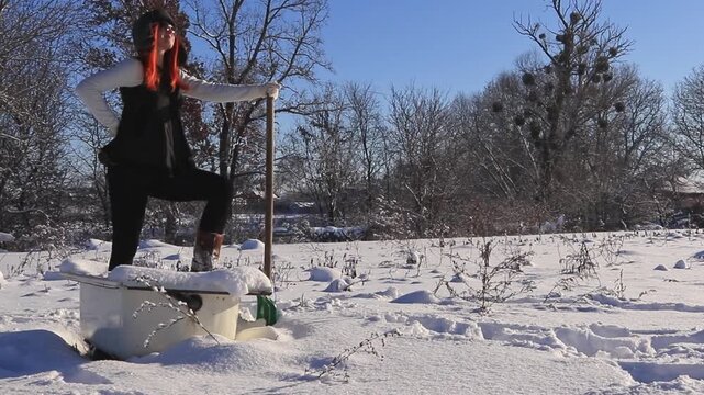 A woman in winter clothes stands in a snowy bathtub in the yard, leaning on a shovel, looking into the distance on a frosty sunny day.