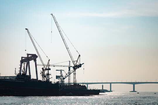 Silhouette heavy industrial cargo vessel sits docked in the harbour while loading containers via port cranes under a sunny sky with sparkling sea waves and a car bridge in Busan, South Korea