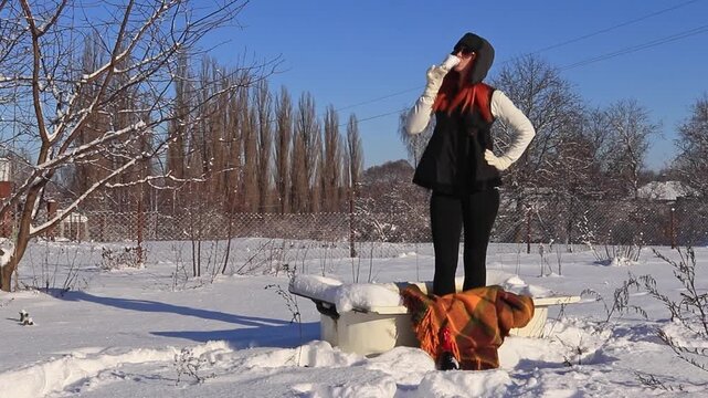 Smiling woman in winter clothes stands with a glass of wine in an abandoned bathtub in a snowy village yard on a sunny frosty day