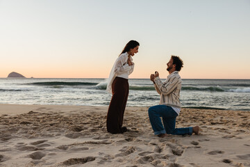 Romantic beach proposal during sunset with man kneeling and presenting ring