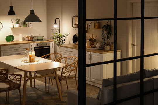 Interior of modern kitchen with glowing lamps, white counters, sofa and dining table at evening