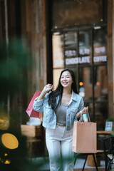 Happy Asian woman holding shopping bags outdoors in a trendy urban area, smiling confidently after shopping, representing modern lifestyle, retail experience, and consumer happiness.