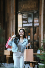 Happy Asian woman holding shopping bags outdoors in a trendy urban area, smiling confidently after shopping, representing modern lifestyle, retail experience, and consumer happiness.