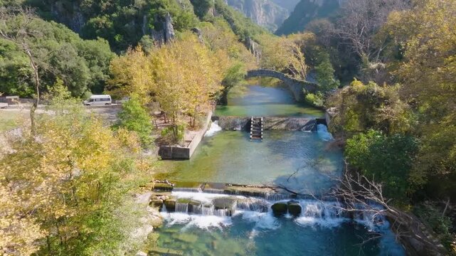 Aerial drone flight over the Voidomatis River, approaching the historic Klidonia stone bridge in Zagori, Greece. Shot reveals the waterfalls, autumn trees, and gorge