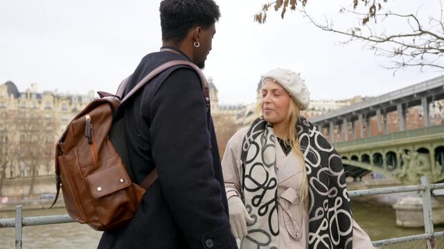 Young and loving multi ethnic couple sharing a pleasant conversation along the seine river in paris, with the bir hakeim bridge in the background, enjoying their european winter vacation together