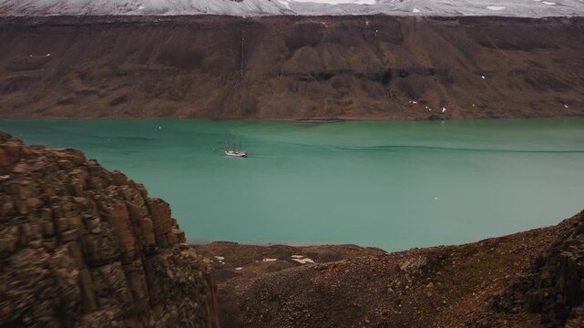 Drone flies over an arctic river flowing down from the alpine into a fjord where a schooner is anchored.