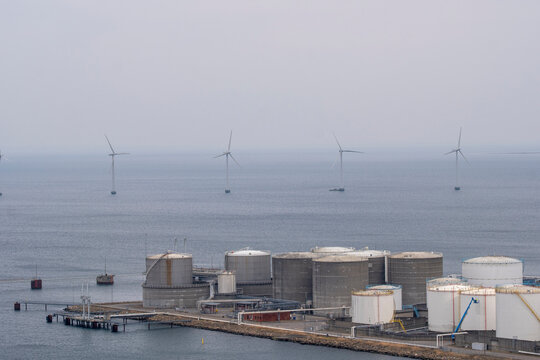 Storage tank terminal on coastline beside sea with offshore wind turbine supports industry energy networks under overcast sky and modern infrastructure