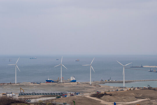 Coastline harbor construction by sea with wind turbine shows renewable industry shipping and broad landscape under overcast industrial mood