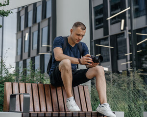 Fototapeta premium Young photographer sitting on a modern wooden bench outside a glass office building, holding a mirrorless camera; casual summer outfit, urban lifestyle portrait.
