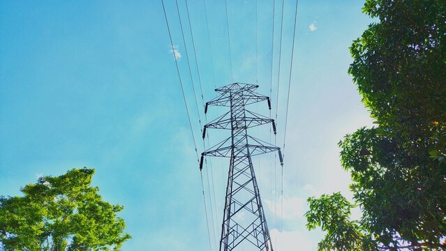 High voltage electricity transmission tower against a clear blue sky surrounded by green trees in the morning.