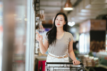 Smiling Asian woman shopping for fresh food in a modern supermarket, placing packaged meat into a shopping cart under bright store lighting, representing everyday grocery shopping lifestyle