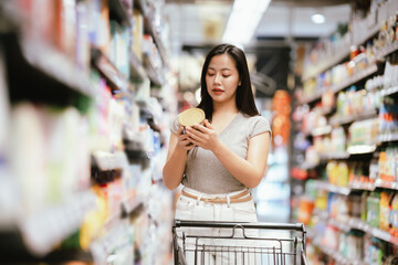 Asian woman reading a food label while shopping in a supermarket aisle, pushing a cart and carefully choosing products, representing mindful consumer behavior and everyday grocery shopping.