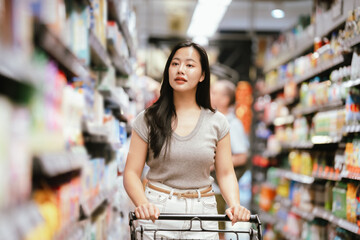 Asian woman reading a food label while shopping in a supermarket aisle, pushing a cart and carefully choosing products, representing mindful consumer behavior and everyday grocery shopping.