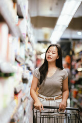 Asian woman reading a food label while shopping in a supermarket aisle, pushing a cart and carefully choosing products, representing mindful consumer behavior and everyday grocery shopping.