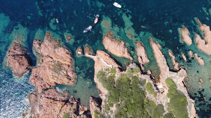 Aerial drone view of a rocky coastline surrounded by crystal clear turquoise sea water with boats anchored near the shore. The shallow transparent water reveals underwater rocks and reef formations, c