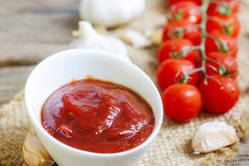 Bowl of rich tomato sauce sits on rustic surface, surrounded by fresh cherry tomatoes and garlic cloves, creating vibrant and appetizing scene
