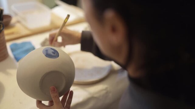 Ceramist applying blue glaze creating decorative patterns on handmade ceramic pottery in a workshop, focusing on creativity and artistic craftsmanship