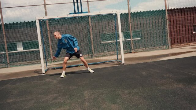 goalkeeper on urban pitch in rain, crouched ready to save incoming shot, wearing blue jersey and gloves, positioned before net and goalpost, asphalt surface with puddles, chainlink fence