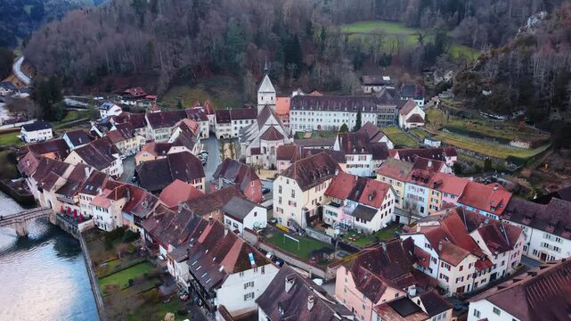 Aerial view of a picturesque town with red-roofed buildings and a church, nestled in a valley with water flowing through, Clos du Doubs, Jura, Switzerland.