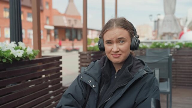 white woman outdoor cafe headphones reacting with playful facial expressions, candid portrait sequence on wooden bench, bundled jacket, potted flowers, urban storefront backdrop, cozy overcast light,