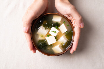 Top view of hand holding healthy Japanese miso soup with soft tofu and seaweed in a wooden bowl on white fabric background